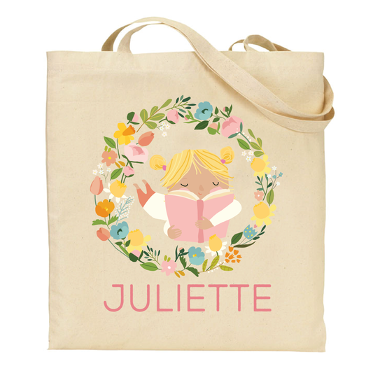canvas tote bag with a design of a blonde girl reading a book surrounded by flowers and the name 'Juliette' on a white background.
